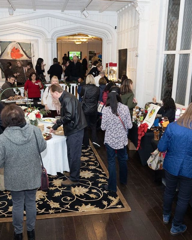 Shoppers crowd the aisles and tables of an indoor holiday market in a hall with tall windows.