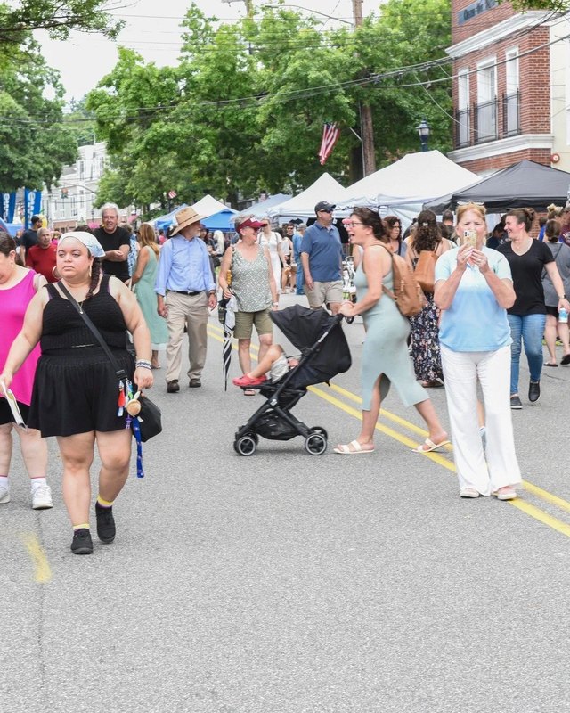 Crowd walking past tents along a tree-lined street in an outdoor market setting.