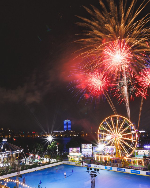 Fireworks burst over a lit Ferris wheel and carousel at a nighttime waterfront festival in Philadelphia, with a bridge in the distance.
