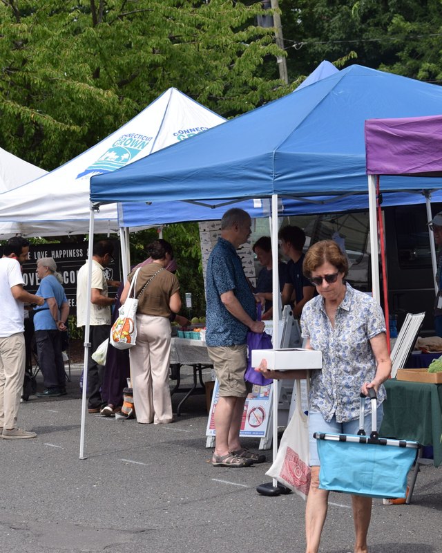 Shoppers browsing produce stands under white and blue market tents at the West Hartford Farmers Market.