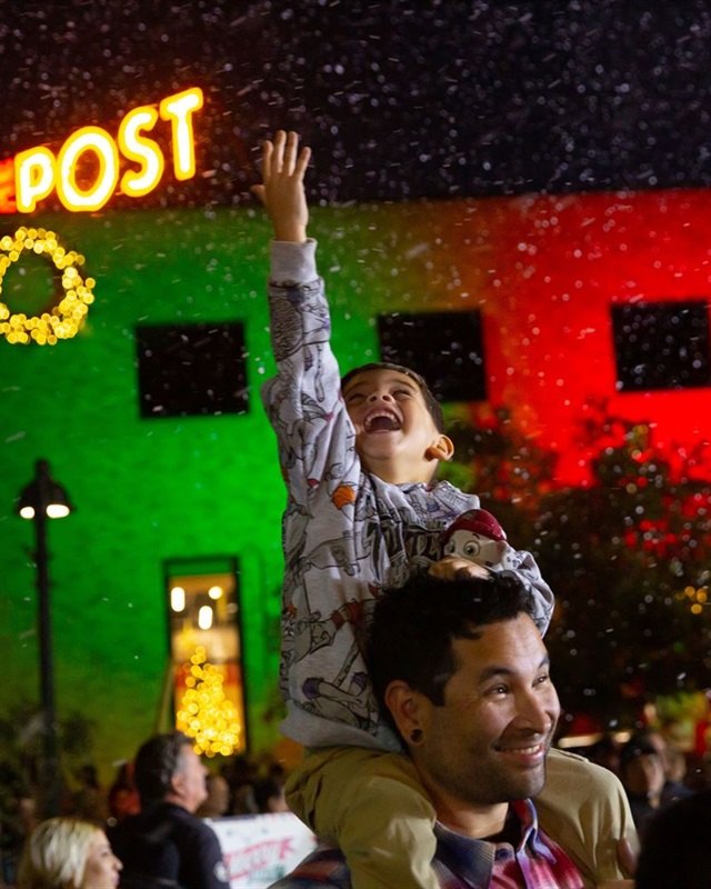 Family with a child raised on shoulders beside a brightly lit holiday building at night, with a crowd below.