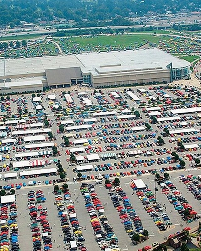 Aerial view of a huge crowded car show lot with rows of parked cars and vendor tents beside a large exhibition hall.