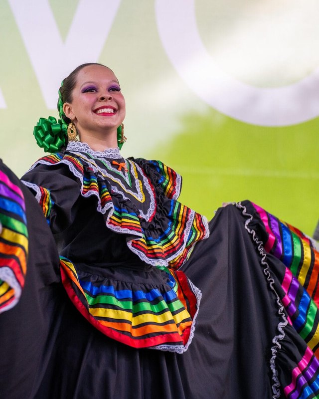Smiling dancer in a black dress with rainbow ruffles performs on an outdoor stage at Avondale Fiesta.