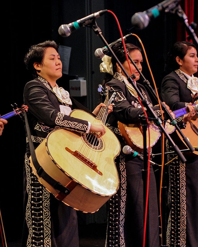 Mariachi musicians in black charro suits with silver embroidery playing guitars and violins on a dark stage.