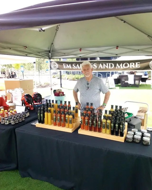 Vendor standing behind a market tent booth stocked with bottles of oil and jars of products