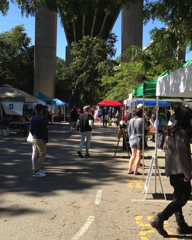 Shoppers walking between market tents at the East Falls Farmers' Market, with concrete bridge supports overhead.