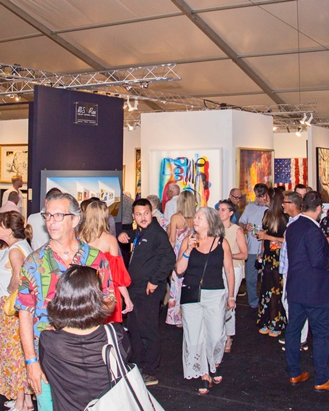 Shoppers and visitors moving through the Hamptons Fine Art Fair tent past large colorful paintings on the walls.