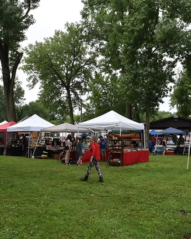 Outdoor market with white and red tents, booths and shoppers spread across a grassy park lawn under tall trees.