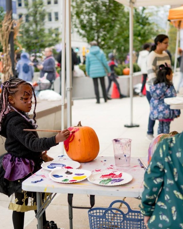 Children painting pumpkins at tables under tents at an outdoor autumn market with vendor booths and shoppers.