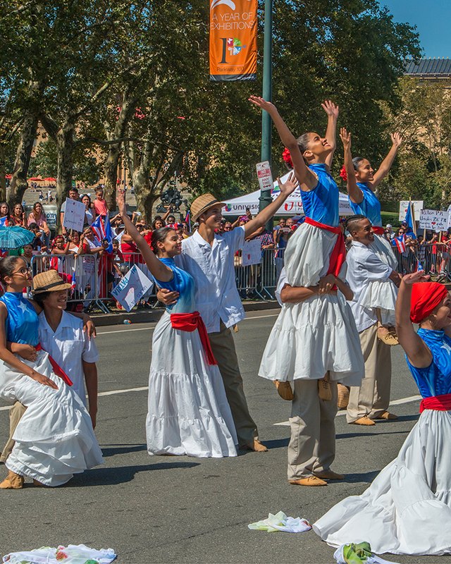 Dancers in white and blue traditional dress performing in the street during a parade crowd