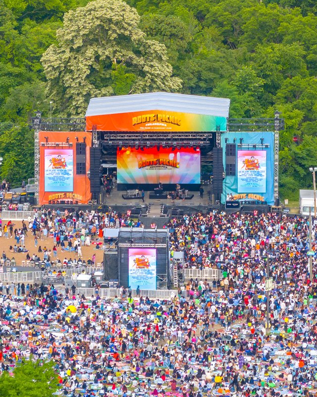 Large crowd gathered before the outdoor stage at the Roots Picnic festival grounds, with trees and tents around.
