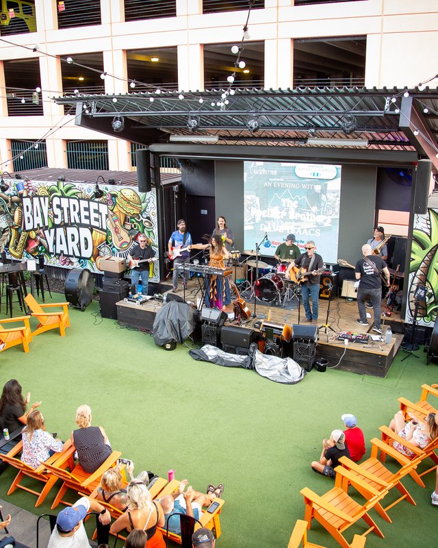 Crowd gathered on turf with orange chairs in front of the Bay Street Yard stage and mural in Fort Myers