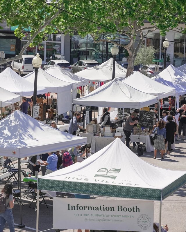 White canopy tents and shoppers at the Rice Village outdoor farmers market, with an information booth in front.