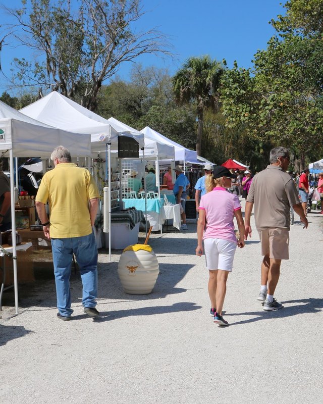Shoppers walking past white canopy booths at an outdoor market, with a honey vendor's table in view.