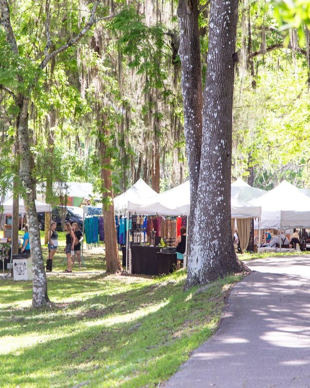 Festival visitors walk among white booth tents under oak trees at Art in the Park in Brooksville.