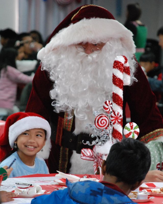 Santa Claus beside children at a holiday breakfast table in a decorated hall, with a child in a red Santa hat smiling.