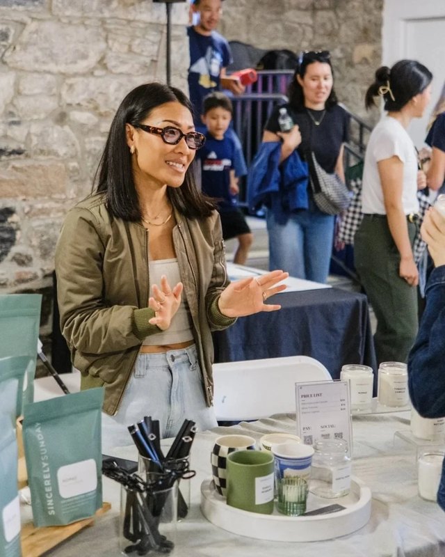 A smiling vendor gestures at her table of mugs and candles, with shoppers behind her in a stone-walled market space.