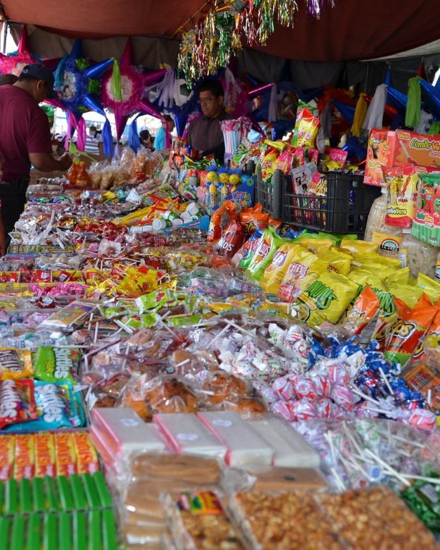 Candy booth with piñatas and bagged sweets, with vendors and shoppers at a market stall.