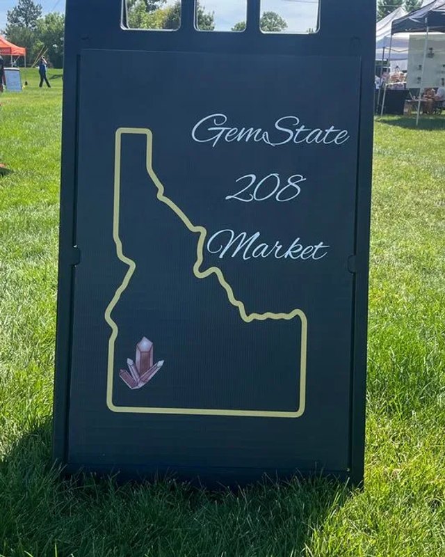 The GemState 208 Market Idaho-outline sign standing on grass, with a few market tents faintly visible in the background.