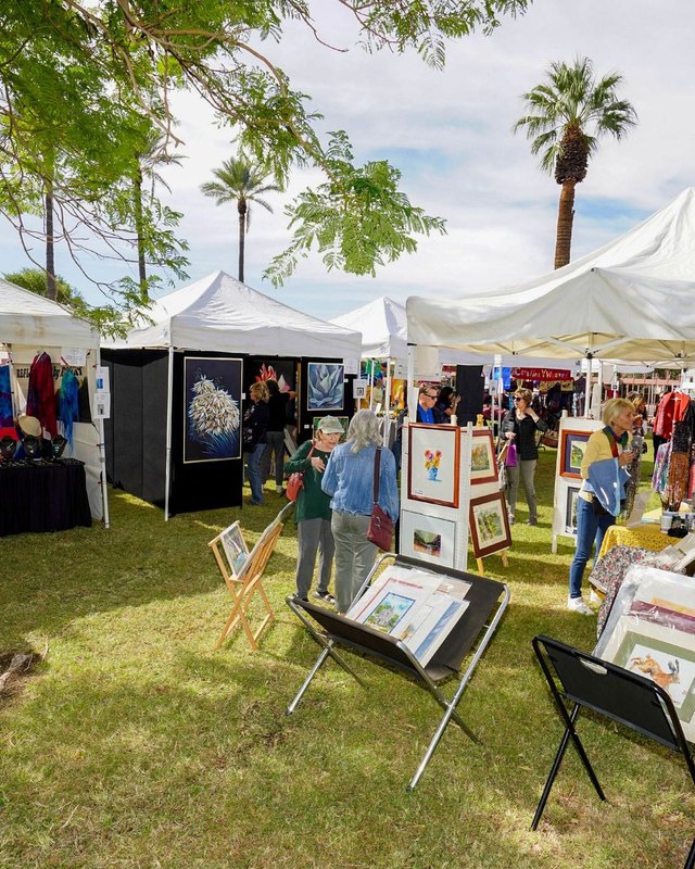 Shoppers browsing white booth tents and artwork displays on grass at an outdoor arts festival.
