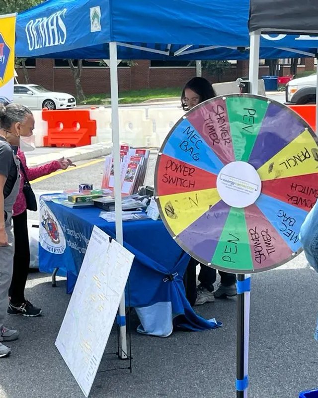 Exhibitor table with a prize wheel under a blue tent at an outdoor community event