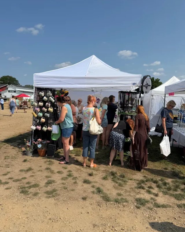 White vendor tent and shoppers browsing at an outdoor fair with a crowd on the grass.