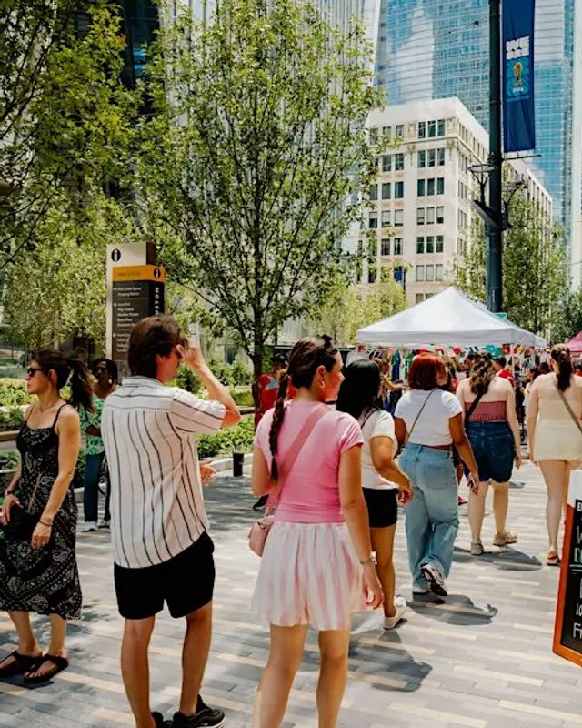 Street crowd walking past white tents and tall green trees on a sunny downtown street, with office towers behind.