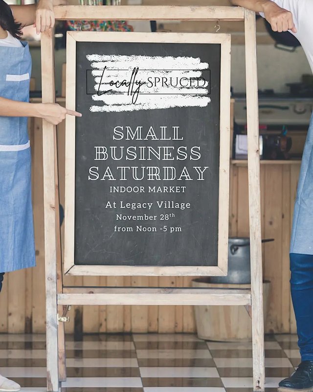 Chalkboard sign advertising the Locally Spruced Small Business Saturday Indoor Market at Legacy Village, held up by two people in aprons.