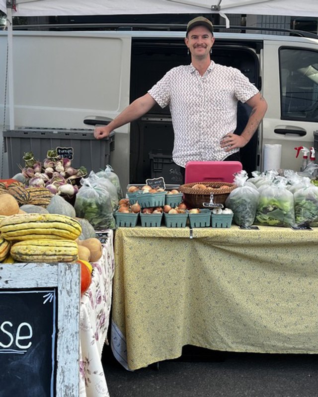 Vendor at Ent Wise Farm standing behind a table of squash and leafy vegetables at the Greeley Farmers' Market.