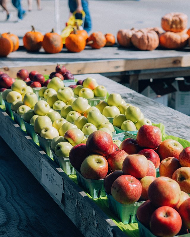 Red and green apples in green cartons on a wooden table at an outdoor market, with pumpkins displayed in the background.