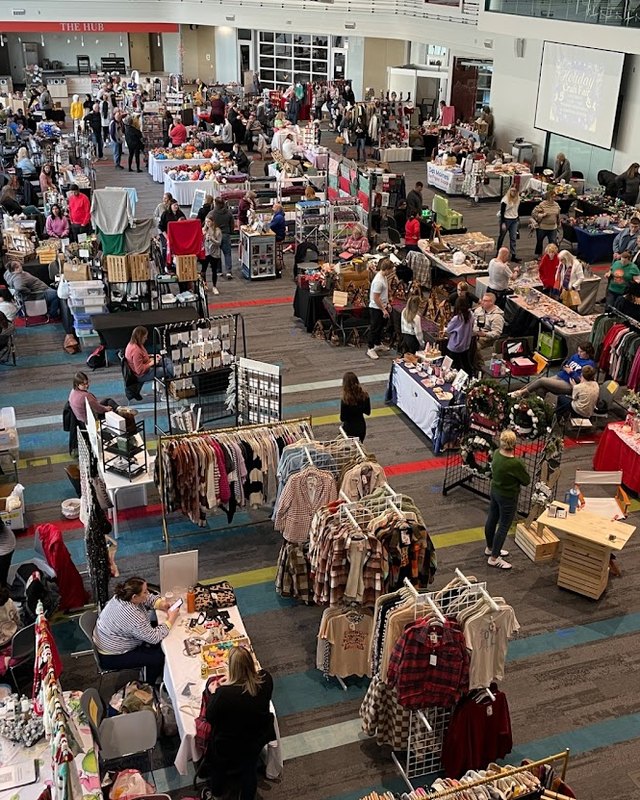 Aerial view of shoppers browsing rows of craft booths at a holiday craft fair in a large hall.