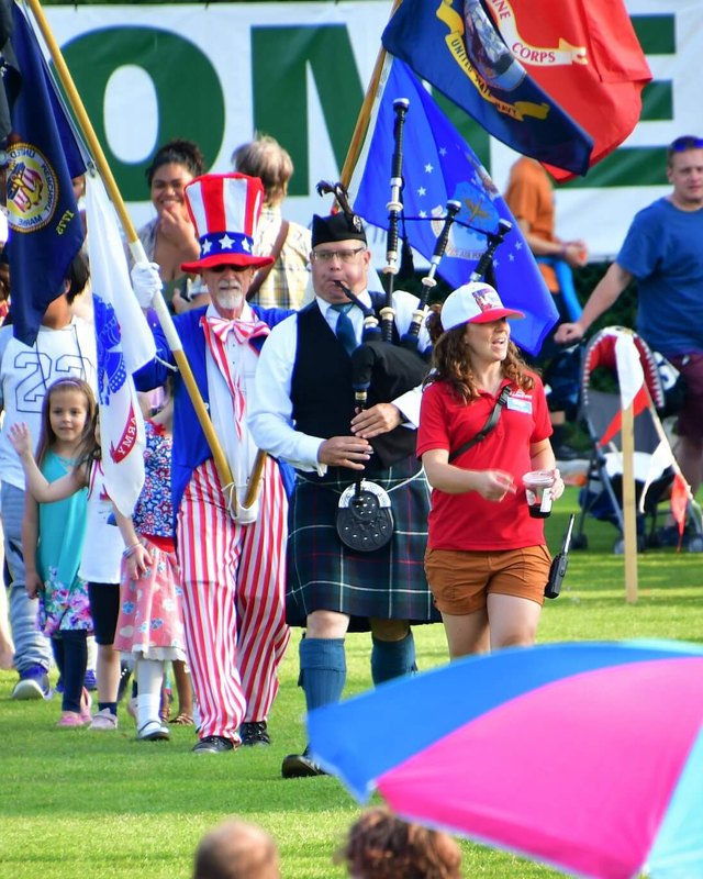 Parade procession on a grass field with a costumed Uncle Sam, a bagpiper, flag bearers and crowds at the Federal Way festival.