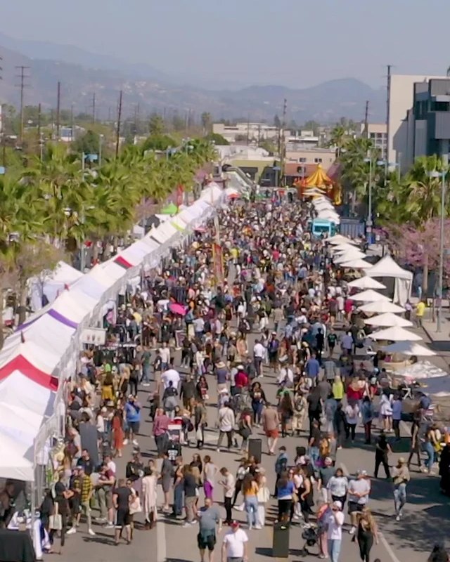 Dense crowd walking down a street lined with white vendor tents at the Vegan Street Fair.