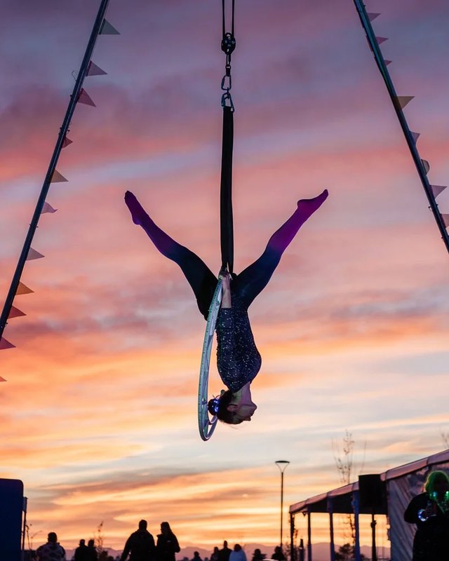 Aerialist hanging upside down from a rig at sunset under festival pennants, with tents and visitors below.