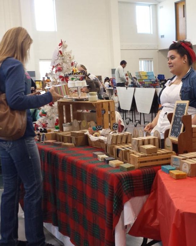 Craft fair vendor booth with wooden handmade items and shoppers inside a bright indoor hall.
