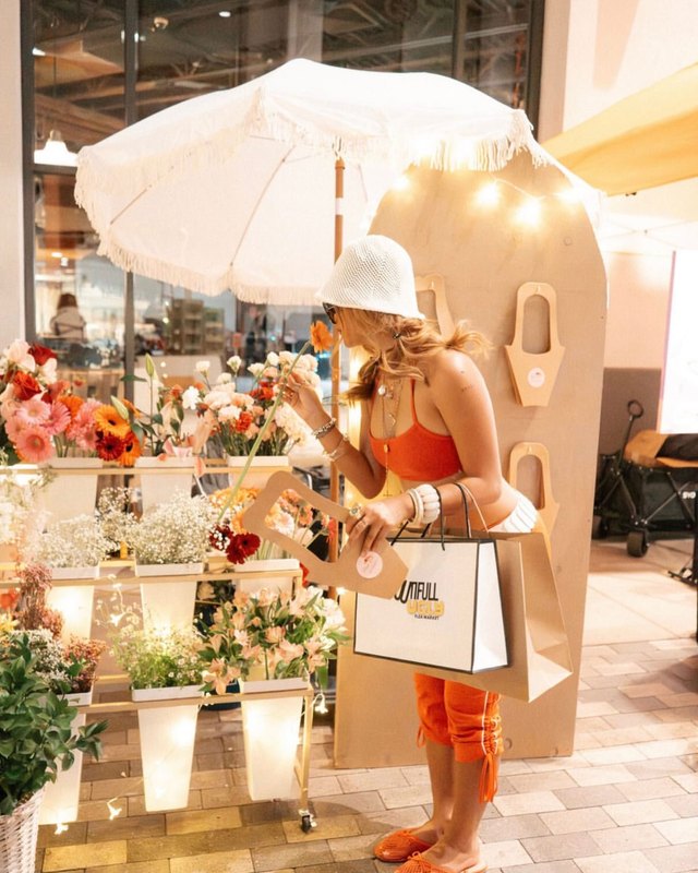 Shopper with a tote bag browsing flower stalls under an umbrella at the Arizona Center market.