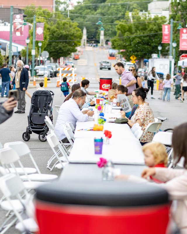 Street fair on a tree-lined street with tents, tables, and shoppers gathered around a long picnic table.