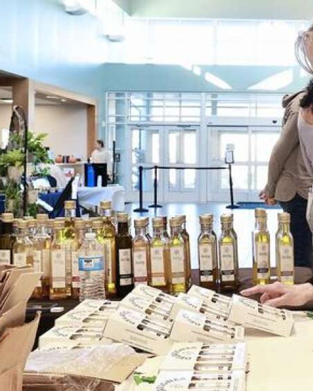 Rows of olive oil bottles and gift boxes on a vendor table in a bright indoor market hall, with a shopper partly visible at the right edge.