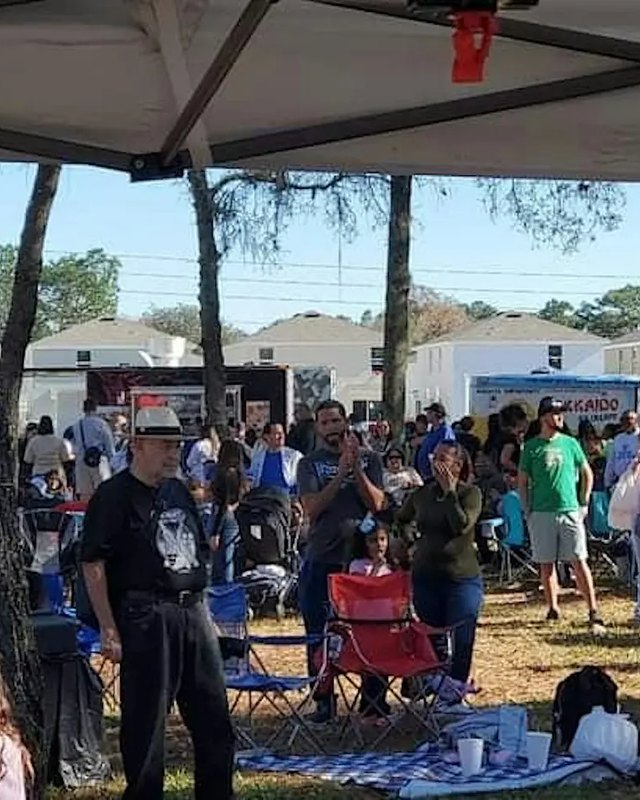 Outdoor festival crowd gathered under white tents and trees, with vendor booths and shoppers.