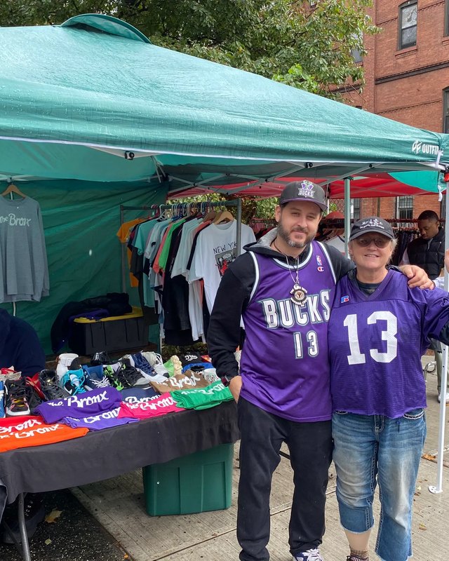 Two men posing in front of a green vendor tent with clothing and goods on tables at the South Bronx Swap Meet.