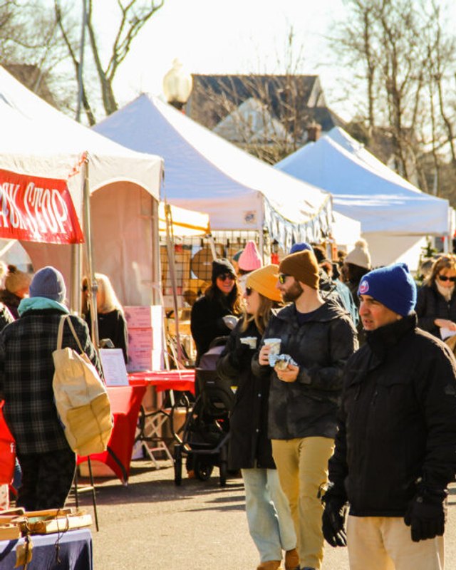 Shoppers browsing holiday market booths with white tents and red-draped tables at an outdoor market.