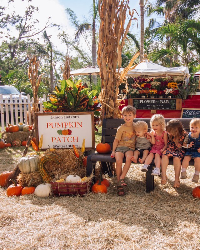 Kids seated on a bench in front of a Pumpkin Patch sign, a red truck and a Flower Bar display, with pumpkins, hay and corn stalks.