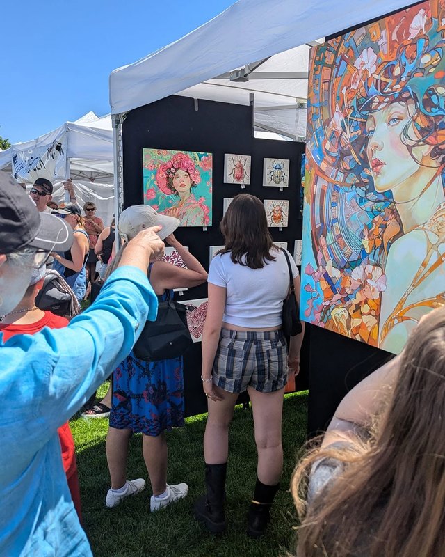 Visitors looking at colorful paintings displayed in a booth at an outdoor art festival.