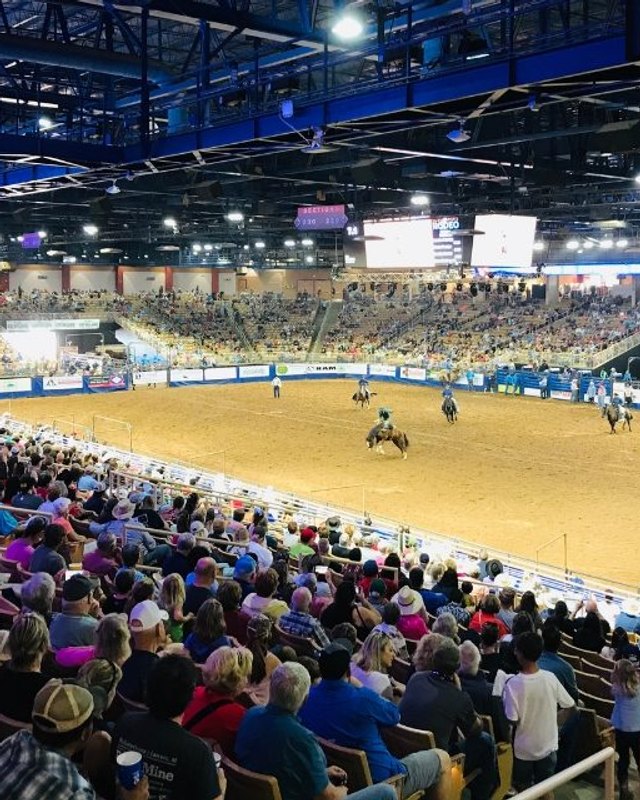Large crowd seated in the stands overlooking an indoor arena during a rodeo event, with riders visible on the dirt floor.