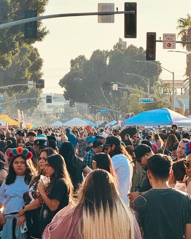 Dense crowd walking past vendor tents on a sunny city street during the Riverside Dia de los Muertos festival.