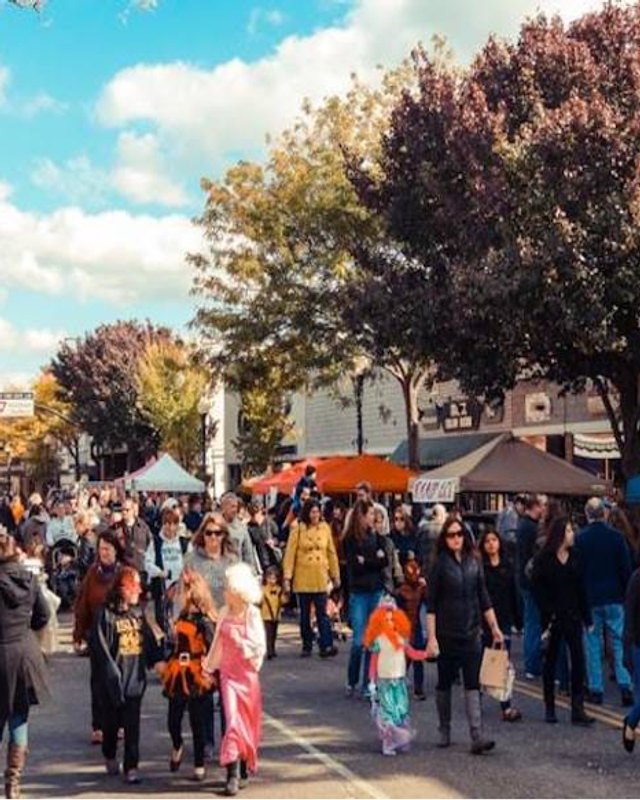Crowds of shoppers browsing vendor tables under autumn trees on a village street.