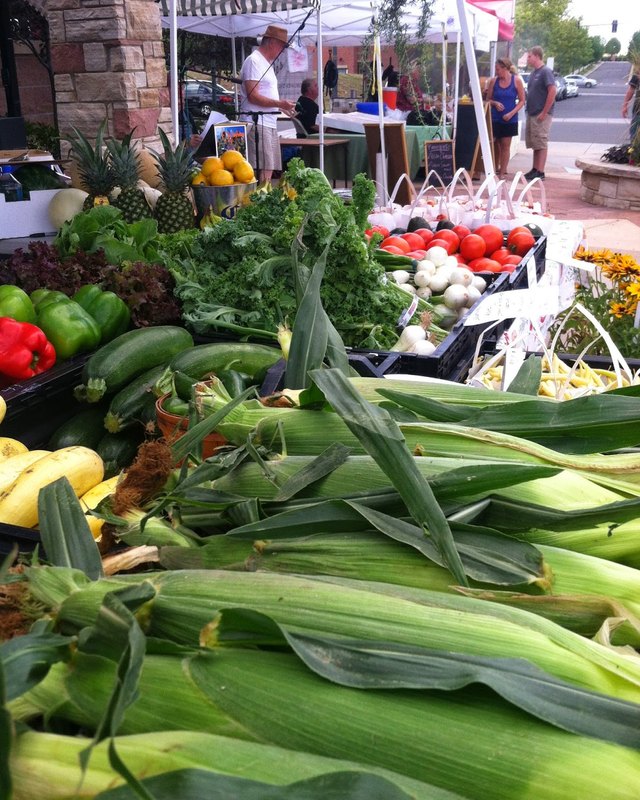 Produce booth with peppers, corn, and squash at an outdoor market with shoppers in the background.