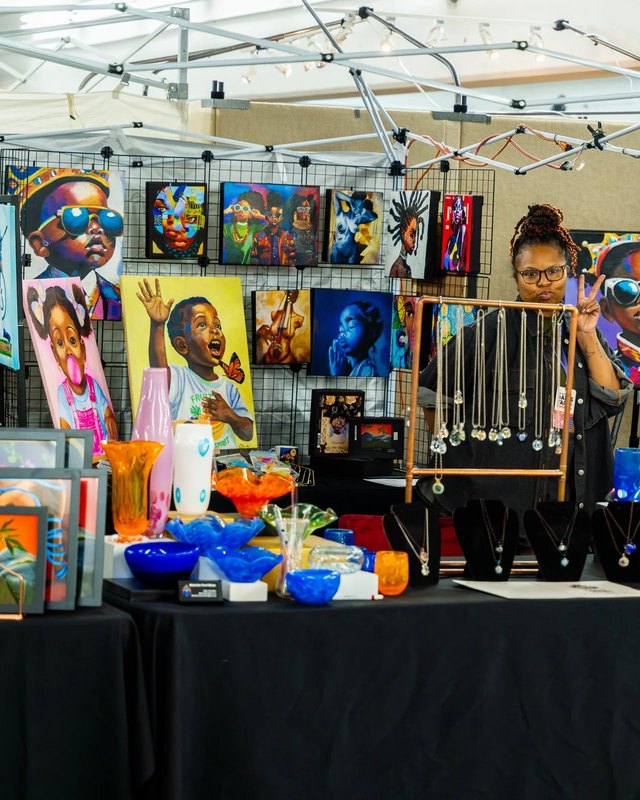 Artist booth at an outdoor fair with colorful paintings, blue and orange glass bowls, jewelry and a vendor behind the table under a white tent canopy.