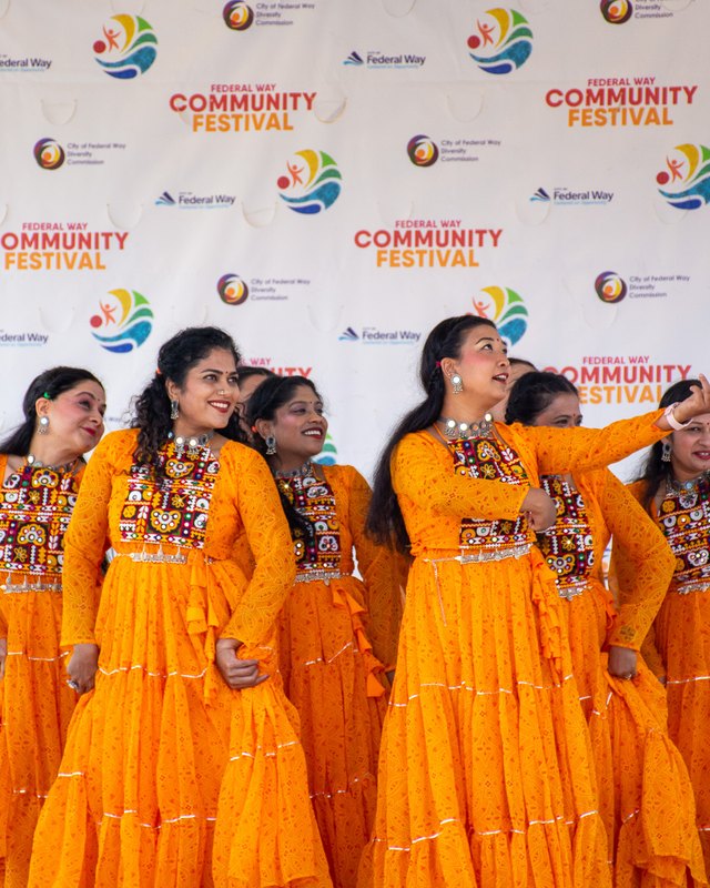 Five women in bright orange traditional dresses pose on stage in front of a Federal Way Community Festival backdrop.