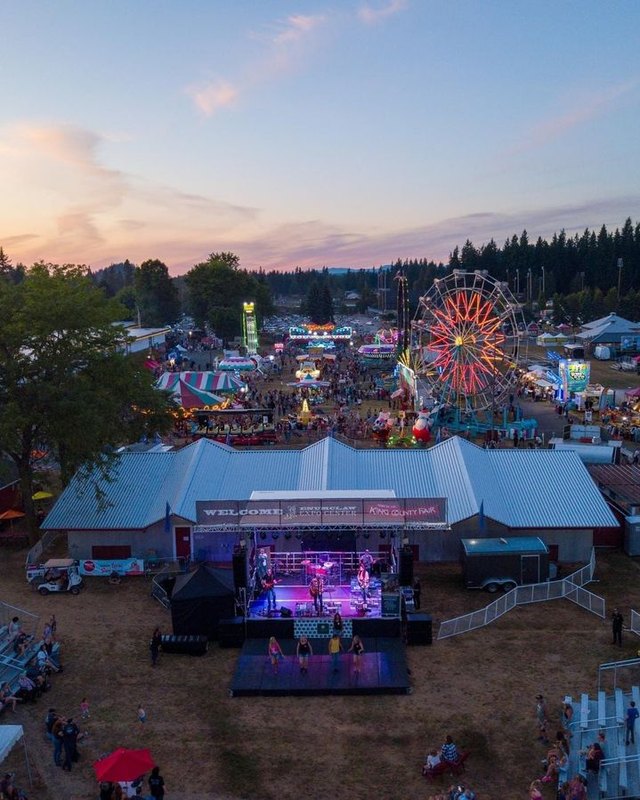 Aerial evening view of the fairgrounds with a lit stage, tents, and a Ferris wheel in the distance.